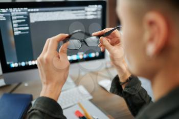 Male IT specialist holds glasses at pc monitor in office. Web programmer or designer at workplace, creative occupation. Modern information technology, corporate team