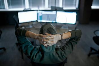 Male internet hacker in hood sitting at screens, back view. Illegal web programmer at workplace, criminal occupation. Data hacking, cyber security
