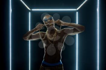 Serious rapper in gold chains poses in illuminated cube, dark background. Hip-hop performer, rap singer, break-dance performing, entertainment lifestyle, breakdancer