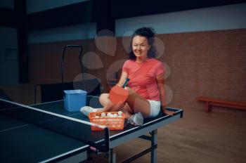Woman sitting on ping pong table, tennis training in gym, female player with basket of balls. Sportive girl playing table-tennis indoors, sport game with racket, active healthy lifestyle