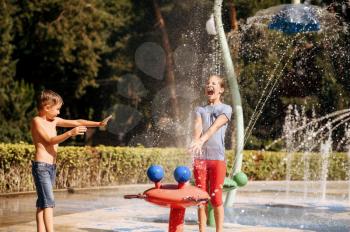 Little boy and girl play with splashes on water playground in summer park. Children leisures in aquapark, aquatic adventure, young brother and sister are having fun in fountain, waterpark
