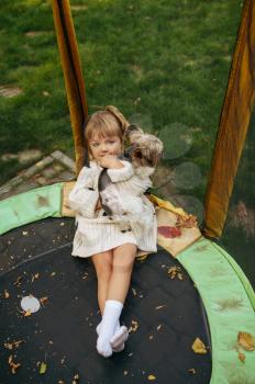 Kid hugs funny dog on trampoline in the garden. Child with puppy leisures on backyard. Little girl and her pet having fun on playground outdoors, happy childhood