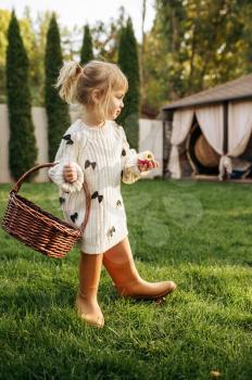 Little girl with basket eats an apple in the garden. Female child poses on the lawn on backyard. Kid having fun on playground outdoors, happy childhood