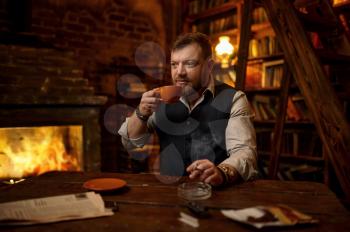 Bearded man with cup of tea smokes a cigarette, bookshelf and rich office interior on background. Tobacco smoking culture, specific flavor. Male smoker leisures