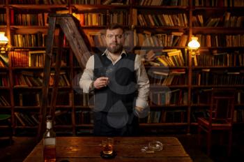 Man smokes a cigar, alcohol beverage in bottle on the table, bookshelf and vintage office interior on background. Tobacco smoking culture, specific flavor. Male smoker leisures