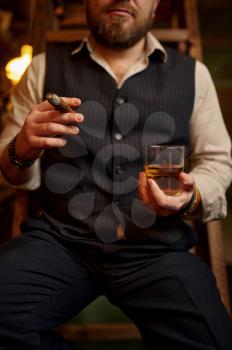 Man smokes a cigar and drinks alcohol beverage, vintage office interior on background. Tobacco smoking culture, specific flavor. Male smoker