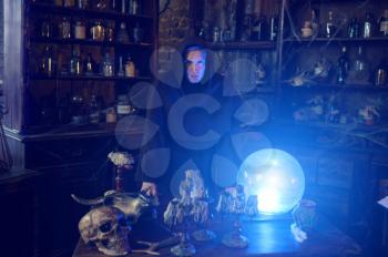 Male exorcist in black hood near the magic crystal ball. Exorcism, mystery paranormal ritual, dark religion, night horror, potions on shelf on background
