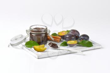 jar of plum jam, fresh plums and cinnamon sticks on wooden cutting board