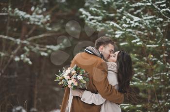 A gentle kiss of the newlyweds in the winter among the dark pine forest.