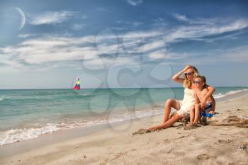blonde mother and son sitting on the beach and watch the sailboat