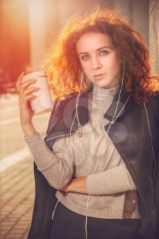 red-haired girl with a glass of coffee is standing by the wall. Portrait at sunset