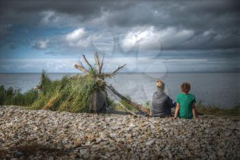 A hut built on the shore by the sea stands. Mother and son are sitting