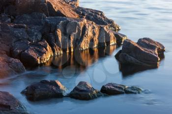 Rocks of stony coast in blue water of lake in yellow morning sun light in sunrise, minimalist nature landscape