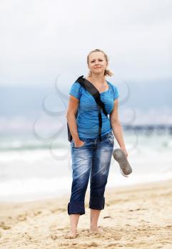 Pretty young blonde woman barefoot with a backpack standing on the beach against the sea. Posing girl outdoors. Selective focus on model. Vertical shot.