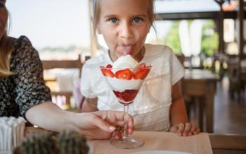 Little girl eating ice cream with strawberries in a restaurant. Tasting the language looking into the camera. Shallow depth of field. Focus on the eyes of the model.