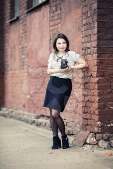 Girl with camera. Pretty young woman with vintage camera in hands posing against brick wall background. Selective focus on girl. Vertical shot. Toned photo with copy space. Vintage style photo.