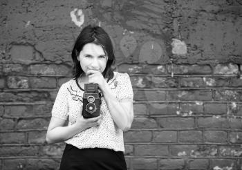 Woman photographer with retro camera in hand on a brick wall background. Girl is thinking and looking at camera. Grayscale photo with copy space.