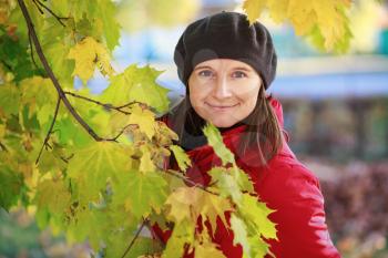 Woman in a beret and red jacket on the background of green and yellow autumn maple leaves. Female portrait. Selective focus on model.