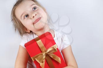 Little girl holding a red gift box with golden ribbon tied bow. Child stuck out his tongue with pleasure. Space for text.
