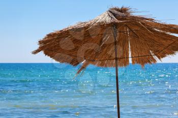 Beach straw umbrella against the bright blue sky and a calm sea. Selective focus on the parasol.