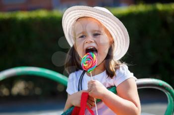 A child in a white hat licks a big colorful lollipop. Shallow depth of field. Focus on the model's face.