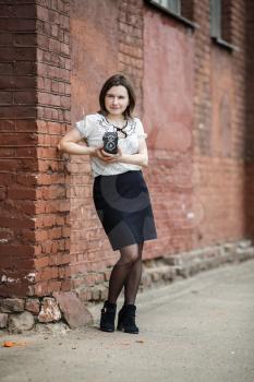 Pretty young woman with an old vintage camera in hand posing on a background of an old brick wall. Shallow depth of field. Selective focus on model. Vertical shot. Retro style photo.