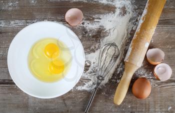 Cooking background with eggs, raw eggs in a dish, eggshells, flour, rolling pin and whisk on a wooden background. Top view.