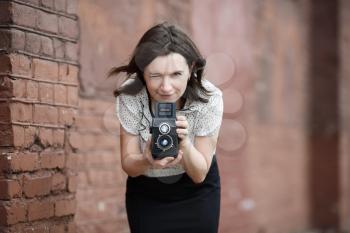 Woman photographer with an old vintage camera in hand on a brick wall background. Pretty young woman taking photo outdoor. Selective focus on the model's face and camera. Retro style photo.