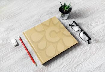 Blank stationery template. Closed blank sketchbook, glasses, pencil, eraser and plant on light wood table background.
