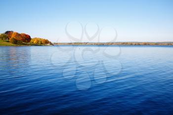 Big lake landscape. Blue sky reflected in calm water. Autumn trees on the shore in the distance.