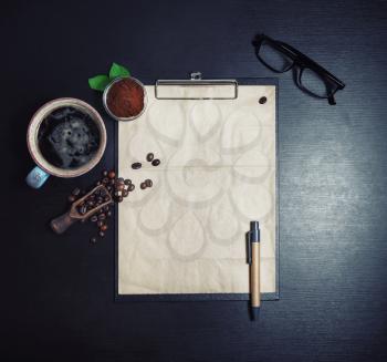 Clipboard with blank kraft sheet of paper, coffee cup, coffee beans, pen, glasses and ground powder on black wooden table background. Template for placing your design. Flat lay.