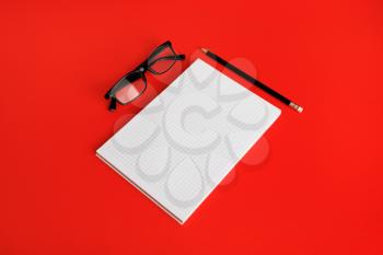 Photo of blank copybook, glasses and pencil on red paper background. Copy space for text.