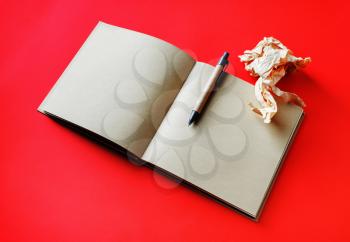 Opened book with blank craft paper pages, pen and crumpled paper on red background.