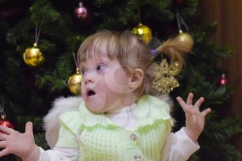 Surprised girl. Child Mimicry. Girl throws up his hands. One year old baby girl sitting on the background of the Christmas tree. A child with gray eyes and blond hair.