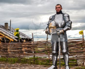 Knight in armor on the background of a wicker fence, a wooden house and the expanses of the motherland. Knightly armor and weapon. Semi - antique photo.