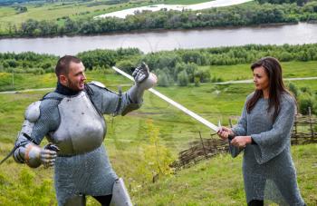 A lady in chain mail and a knight in armor learn to hold a weapon and defend themselves from the blows of a sword. Knightly armor and weapon. Semi - antique photo.