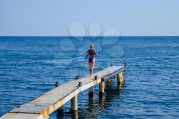 A beautiful girl in a blue bikini walks the pier to the sea. Marine concrete pier. Jumping into the water from the pier. Beautiful Booty Girls.
