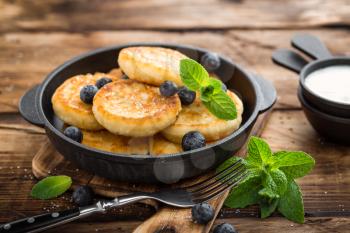 Delicious cottage cheese pancakes or syrniki with fresh blueberry in cast-iron pan on dark wooden rustic background. Tasty breakfast.