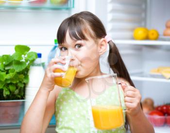 Happy girl drink orange juice standing near refrigerator with fruits and vegetables