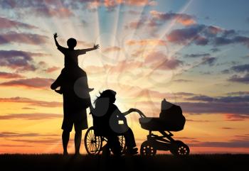 Disabled woman in wheelchair holding baby carriage and beside her husband holding baby on shoulders on sunset. Concept disabled and family