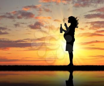 Concept of pregnancy and bad habits. Silhouette of a pregnant woman smokes a cigarette with an alcoholic drink at sunset