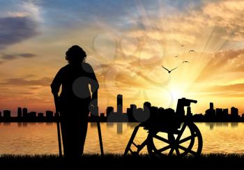 Silhouette of an old woman on crutches on a background of sea sunset urban and wheelchair. Concept of disability and old age