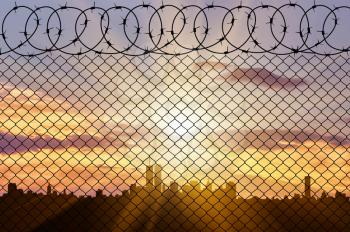 Concept of border service. Silhouette of a border fence with barbed wire on the background of the city at sunset