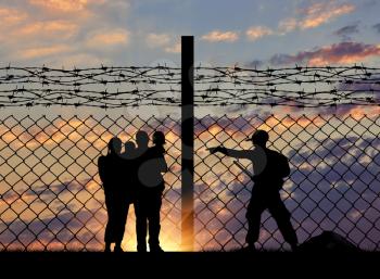 The concept of security. Silhouette of a family with children of refugees and border guards and a fence with barbed wire on the background of evening city away