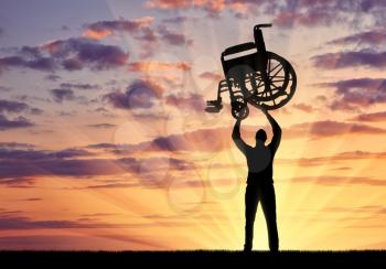 Silhouette of a disabled man standing while lifting a wheelchair against the sunset background. The concept of people defeating disease
