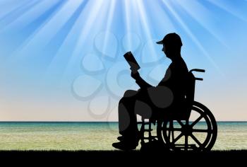 Silhouette of a disabled man in a wheelchair reading a book by the sea. Concept of the way of life of people with disabilities
