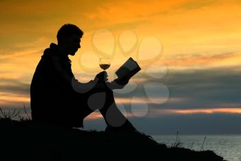 Silhouette guy sitting on breakwater in evening near sea, reads book and drink wine