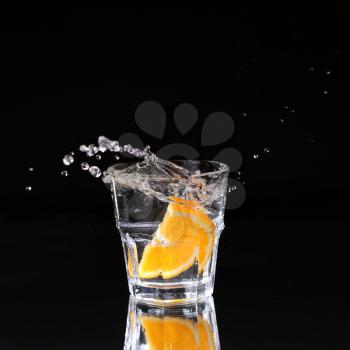 Half of orange falling down in glass with water on deep black. Beautiful glass with sparkling water or other transparent drink and a slice of lemon on black background