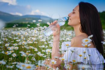 Beautiful woman enjoying daisy field, pretty girl relaxing outdoor, having fun, holding plant, happy young lady and spring green nature, harmony concept. Drinks crystal clear water from a bottle