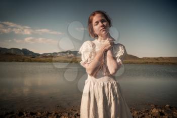 Beautiful young blond woman outdoors portrait near the lake. Red-haired girl with freckles in a white dress. The idea and concept of freedom and loneliness.